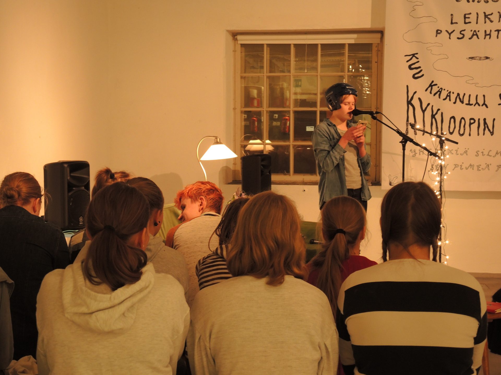 Yleisöä kuuntelemassa open mic -tapahtuman esiintyjää, jolla on jääkiekkokypärä päässään. Esiintyjä lukee esitystään älypuhelimelta. Taustalla näkyy suurikokoinen sanataideteos.