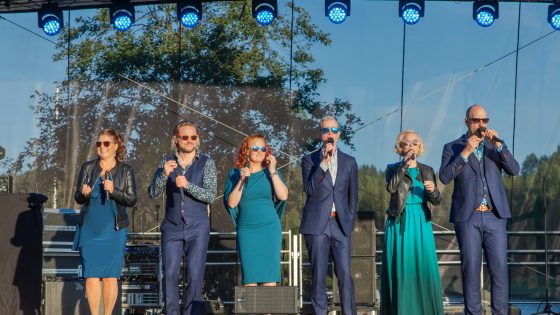 Kuuden laulajan ryhmä lavalla aurinkoisena kesäpäivänä. Kolmella laulajista on tummahkon sinivihreät, erilaiset mekot, ja kolmella laulajista puku tai kauluspaita ja liivi puvunhousujen kanssa. Kaikilla laulajista on aurinkolasit, ja osa heistä hymyilee toisten laulaessa.
